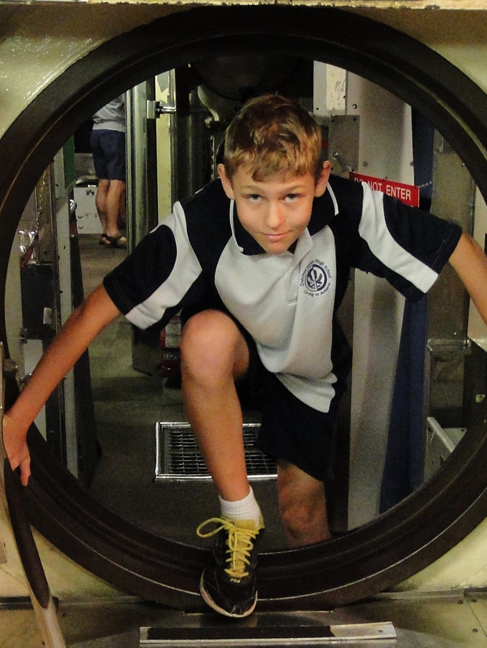 A boy dressed in school uniform climbs through a small hatch door in a submarine