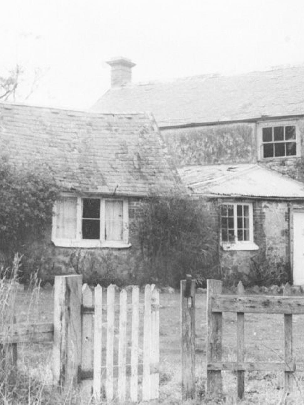 A black and white photograph of an old farmhouse and surrounding fence