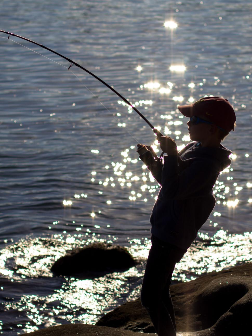 A young boy wearing a hat casts out to sea with sunlight sparkling on the water behind him.