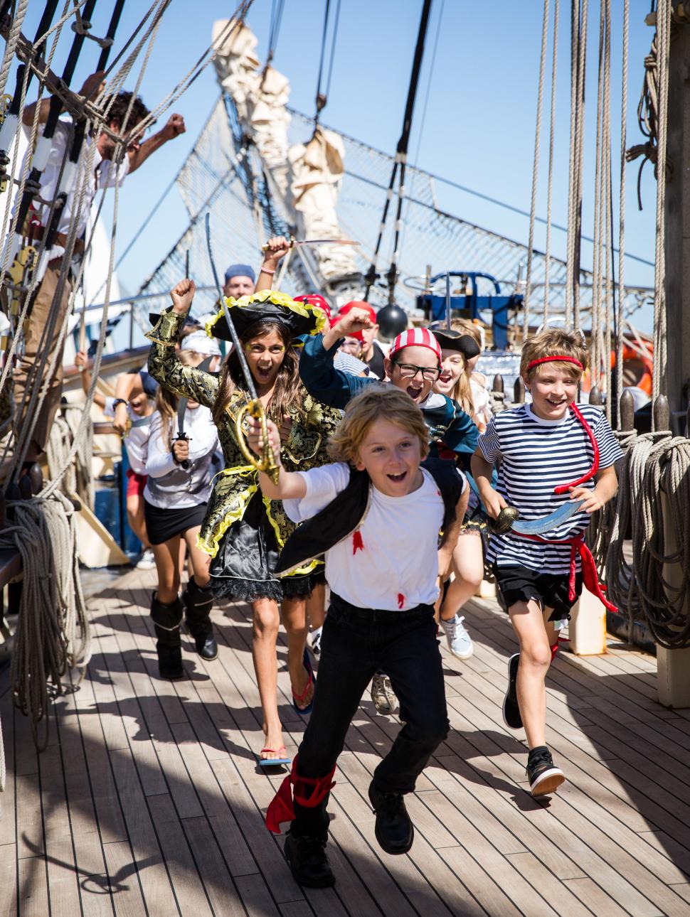 A group of children dressed as pirates rush along the desk of a sailing ship, shouting and waving toy swords