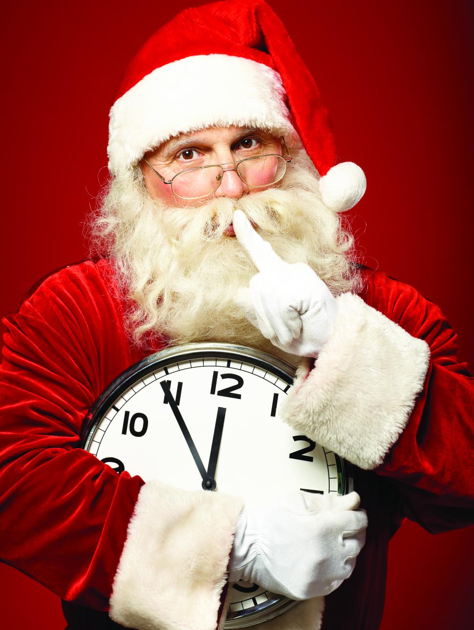 A Santa Claus holding a large clock holds one finger to his lips in a secretive shushing gesture