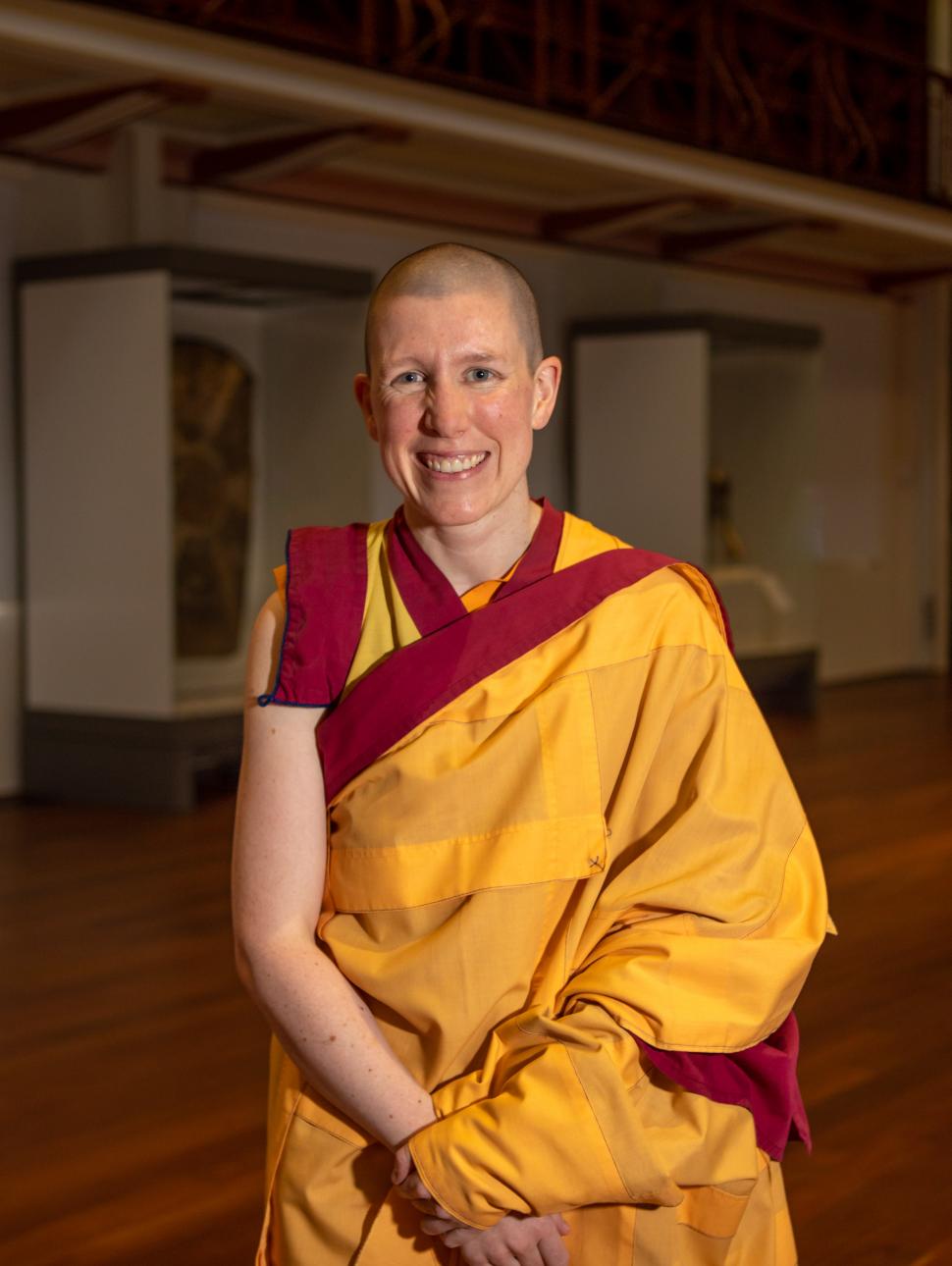 A female monk wearing a yellow wrap garment sits smiling in a hall surrounded by wooden features (Hackett Hall, located at WA Museum Boola Bardip)