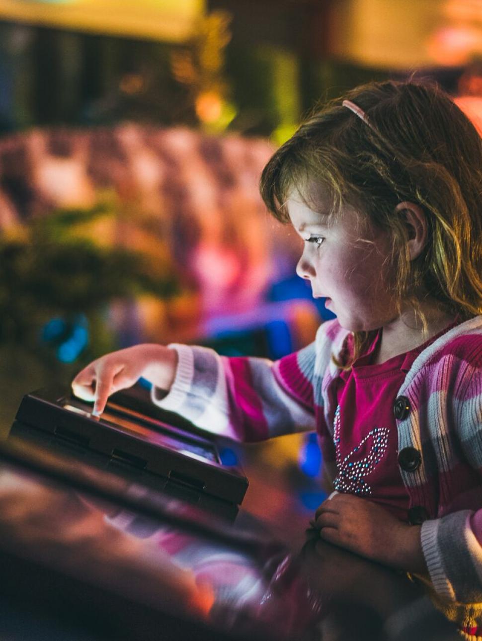 A small girl wearing a pink hooded jumper interacts with a screen about dinosaurs, while one sits blurred in the background behind her