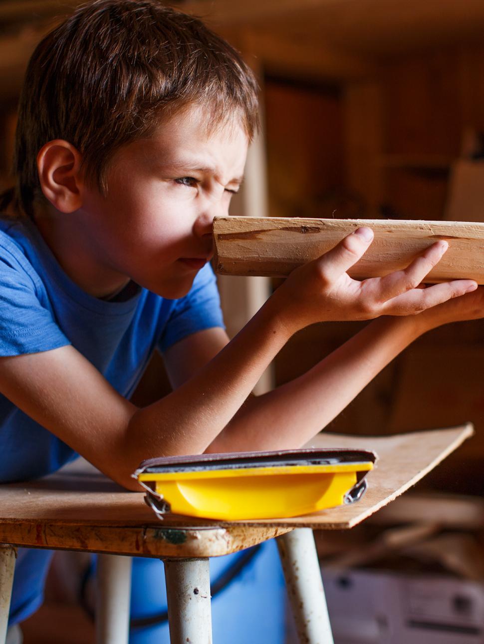 a young boy with brown hair holds a small wooden ship to his face, looking along it