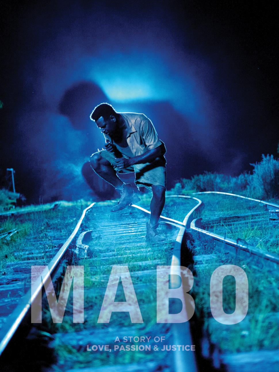 Pictured is a blue tined image of a man in a shirt and shorts with one leg lifted up and head tilted downward with hands clenched close to their chest. They are standing in between two train tracks as the tracks converge. It is dark/night and there is a bright light behind the person 