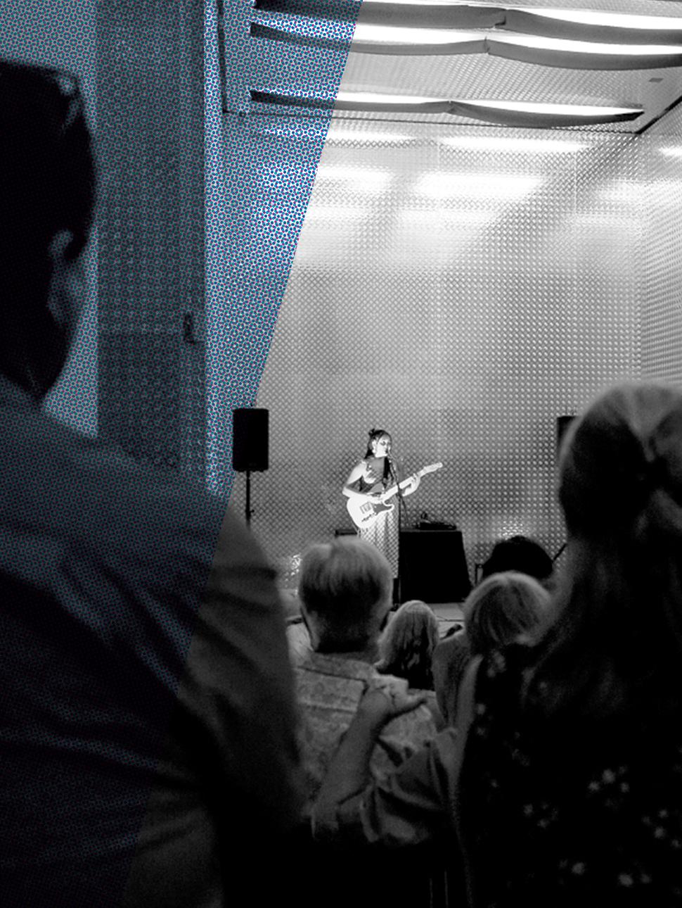 A lone singer stands in the lift entrance with a guitar, a blue strip running along the left side of the black and white image