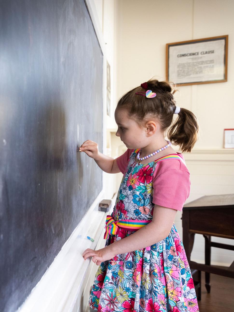 A girl in a pink dress stands at a black chalkboard to draw