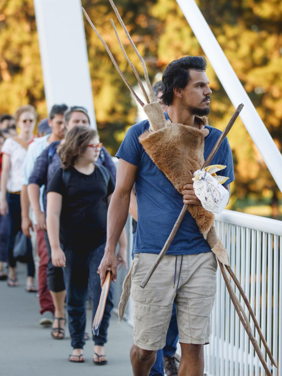 Pictured is a Noongar performer Ian Wilkes holding cultural objects, gazing to the side, leading a group of people single file across a bridge.