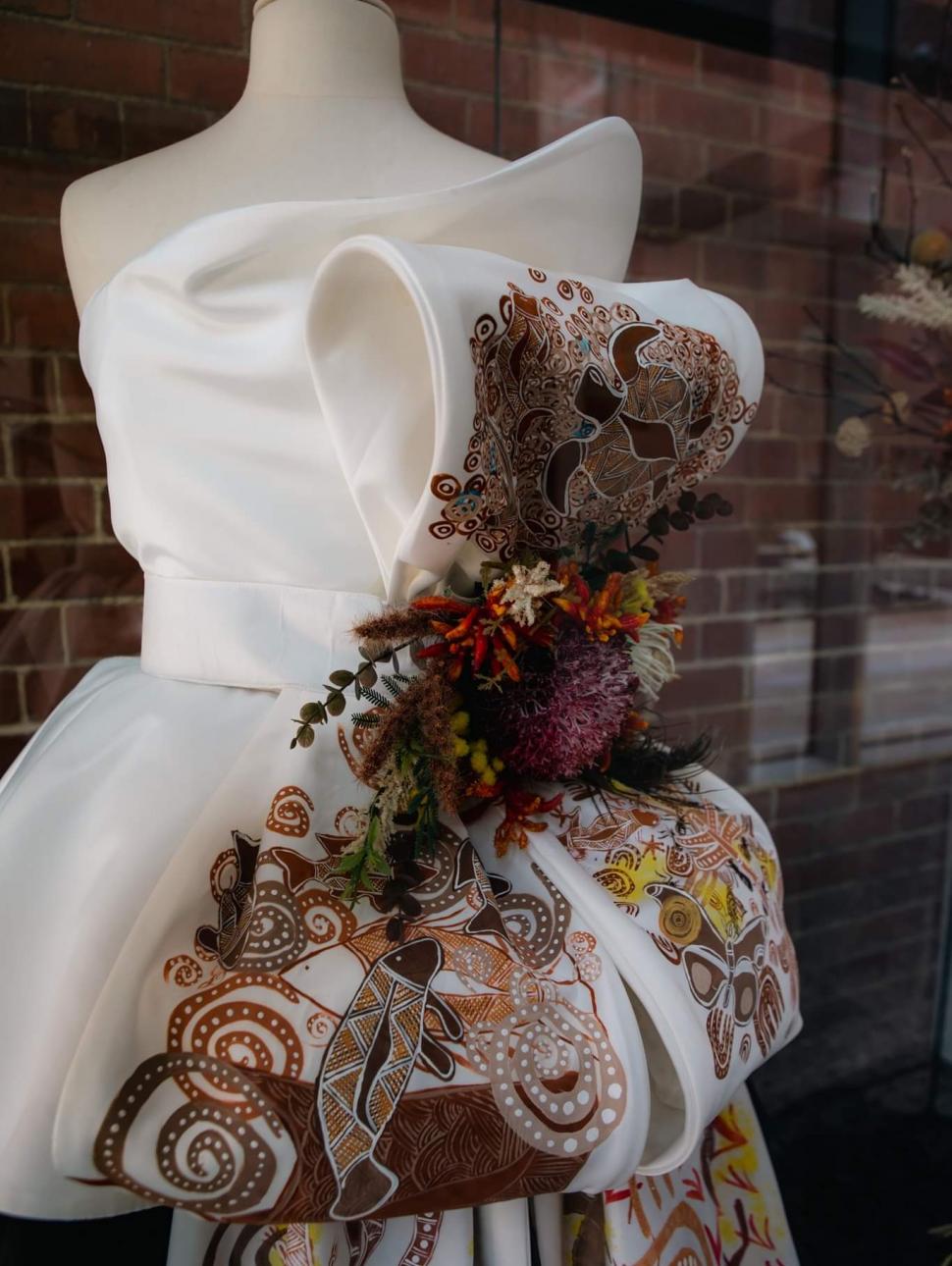 The image shows a haute couture dress made of white materials with a sash encompassing Indigenous designs and wild flowers