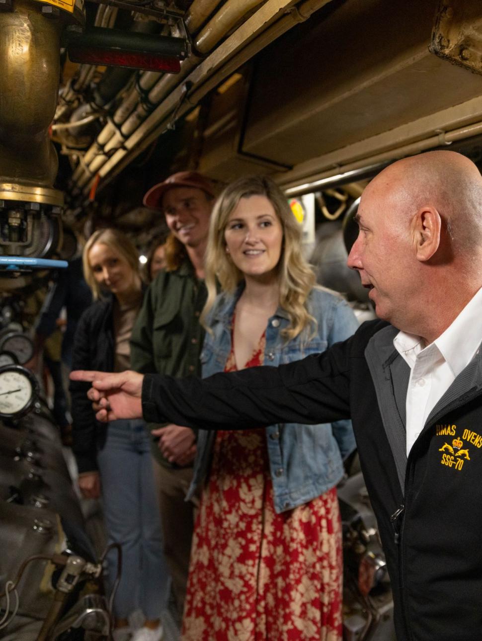 Submarine tour guide gestures inside submarine, three people look on