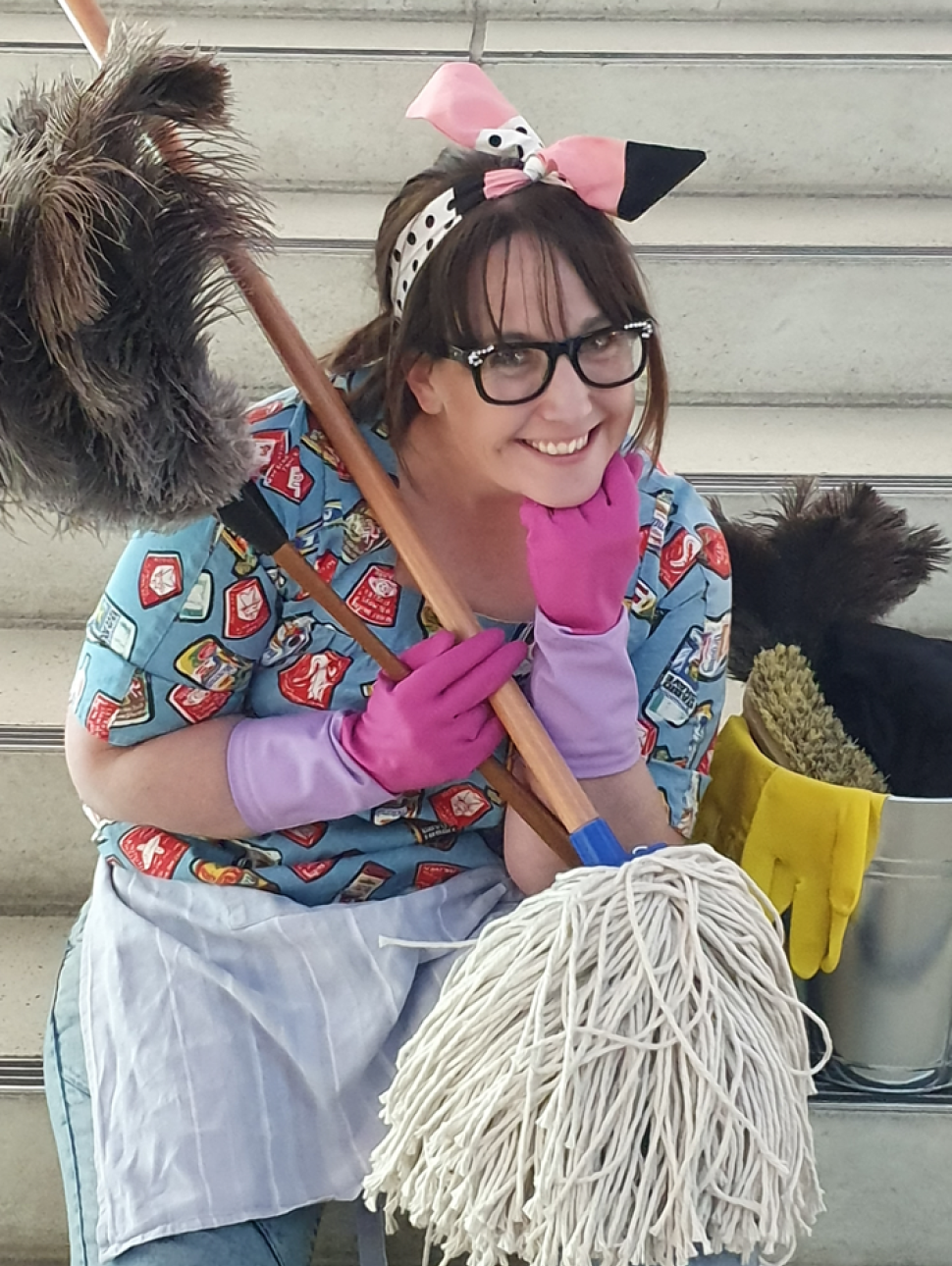 A woman crouches, smiling, while holding cleaning equipment. She wears glasses and pink cleaning gloves, with brown hair pulled back and a ribbon around her head holding her hair in place. 