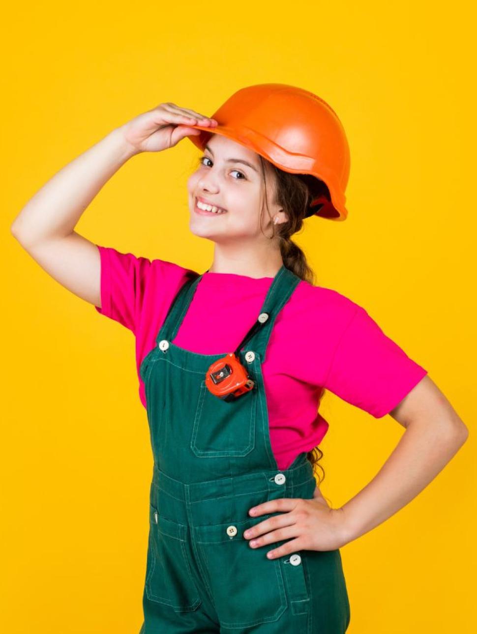 Little girl dressed as a construction worker with tape measure wearnig a orange helmet, pink shirt, green overalls infront of a yellow background. 