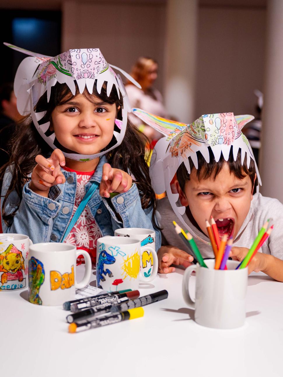 Image of two children roaring at the camera wearing paper dinosaur hats