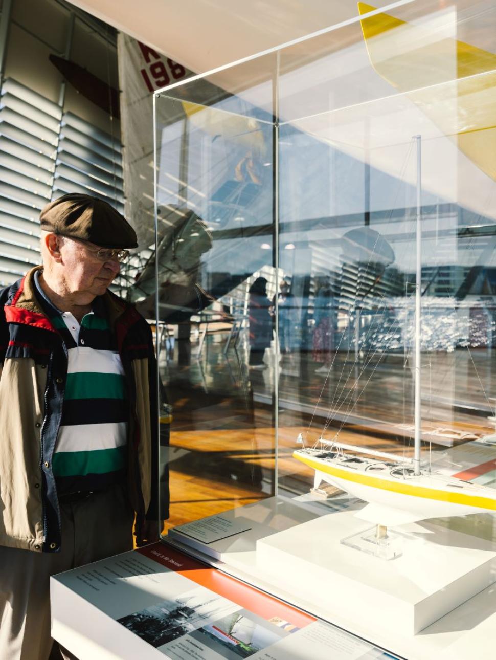 Old gent in hat stairs into glass case holding small boat figurine