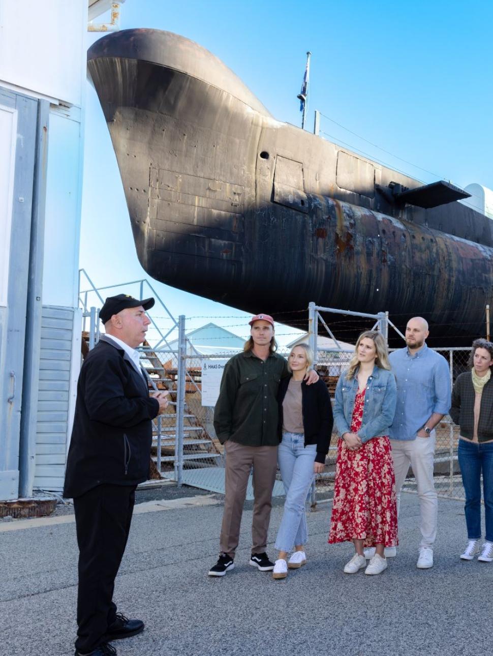 A submarine volunteer speaks to a group of visitors in a semi circle, standing in front of the submarine HMAS ovens