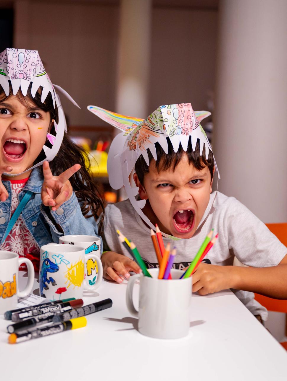 Image of two children roaring at the camera wearing paper dinosaur hats