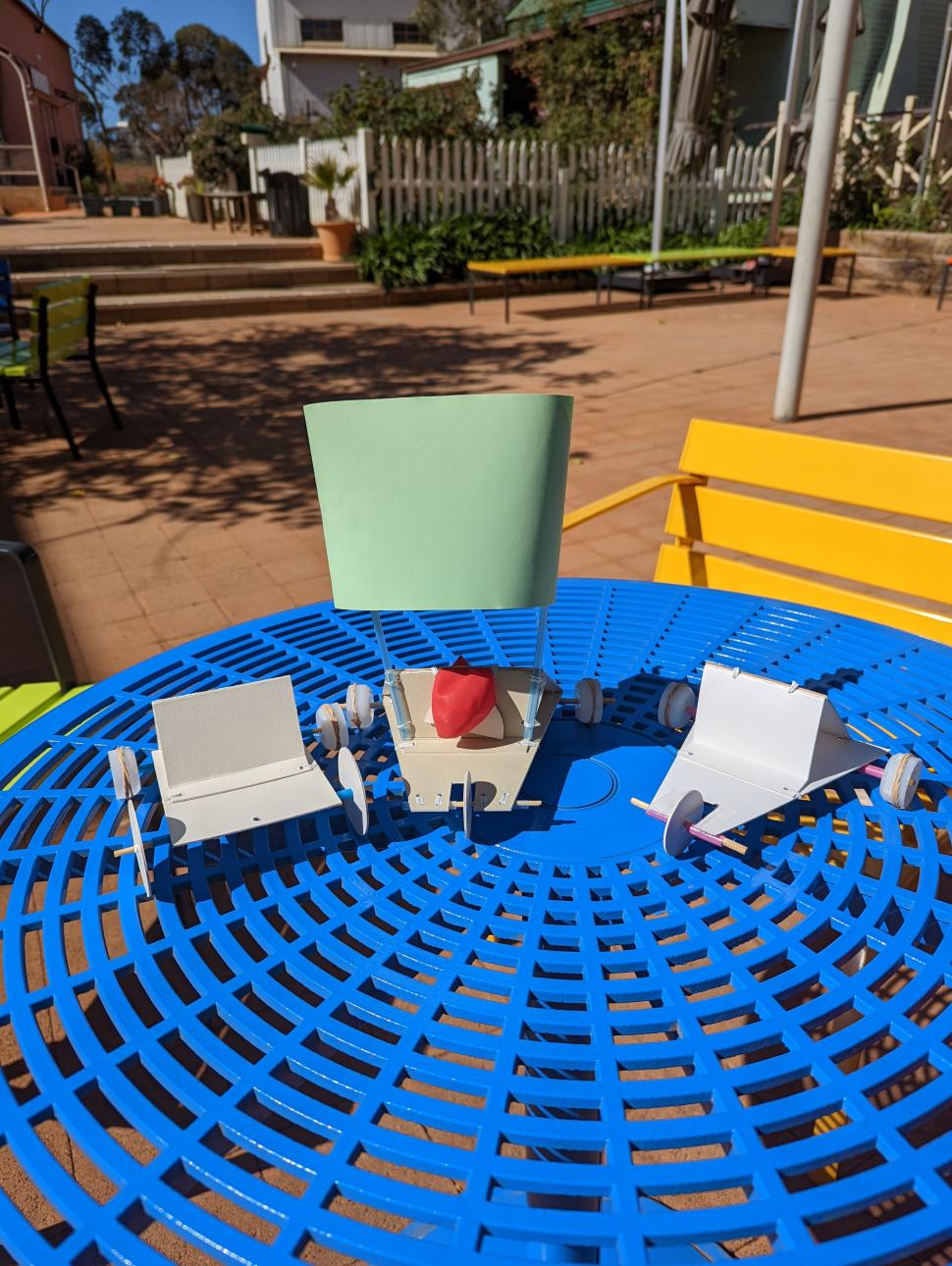 Three handbuilt miniture cars made with recycled materials sitting on a blue bench outside with red brick, benches and a picket fence in the background.