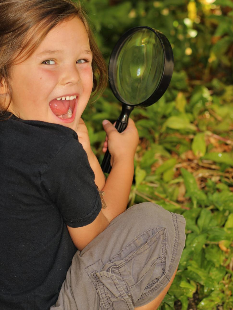 Image of a child in a black t-shirt and grey shorts kneeling, holding a magnifine glass looking back at the camera with greenery in the background.