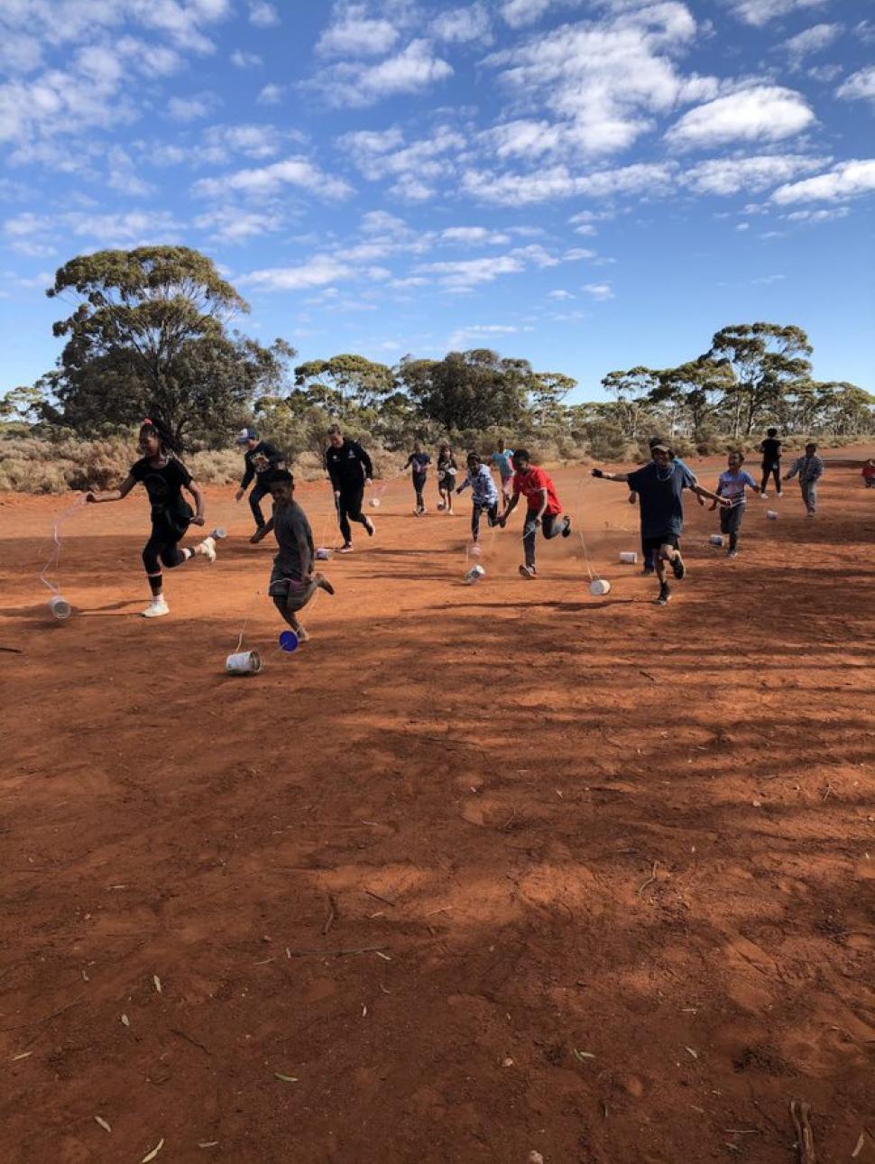 Kids running along a red dirt road playing wth homemade tin trucks made from cans with a blue coloudy sky in the background