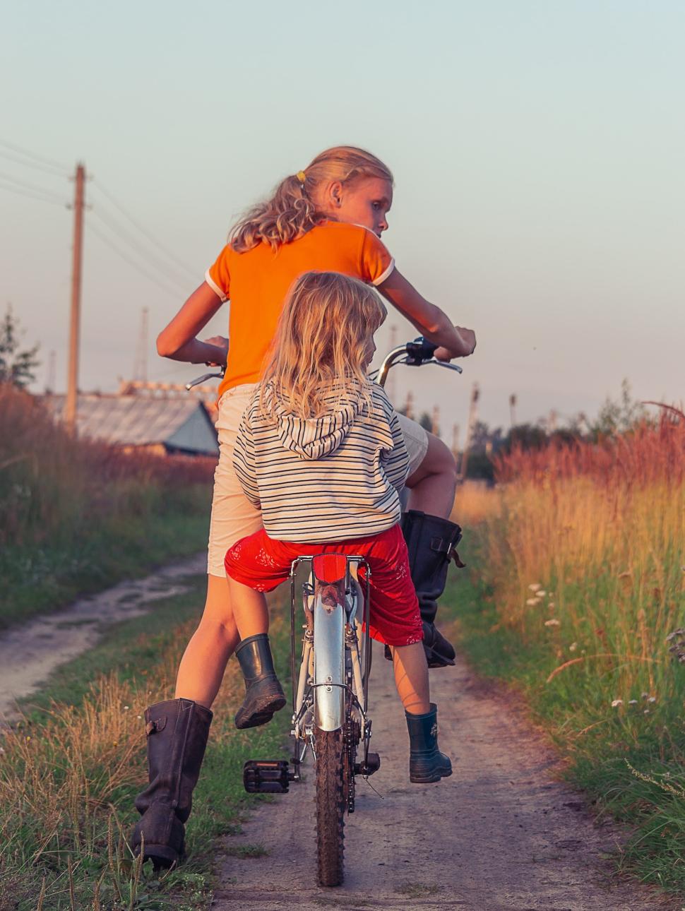 Two young girls riding a bike together facing away from the camera on a dirst footpath in a grassy feild.