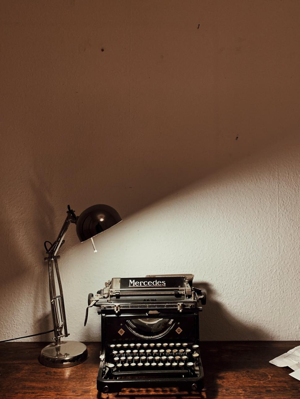 Old typewriter sitting on a wooden bench with a lamp shining on the left-hand corner against a cream wall in dim light. 