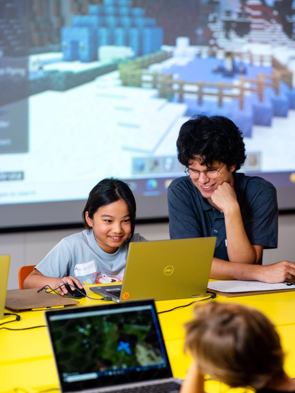 Small girl smiles at laptop on yellow desk, older staff member helps