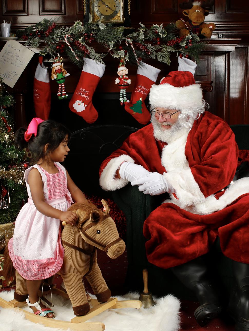 Image of a young girl rocking on a wooden horse looking at Santa Claus sitting on a chair with a christmas tree surrounded by presents and wooden wall in the background
