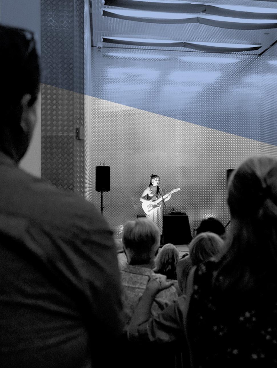 Woman standing in a lift, singing into a microphone holding a guitar