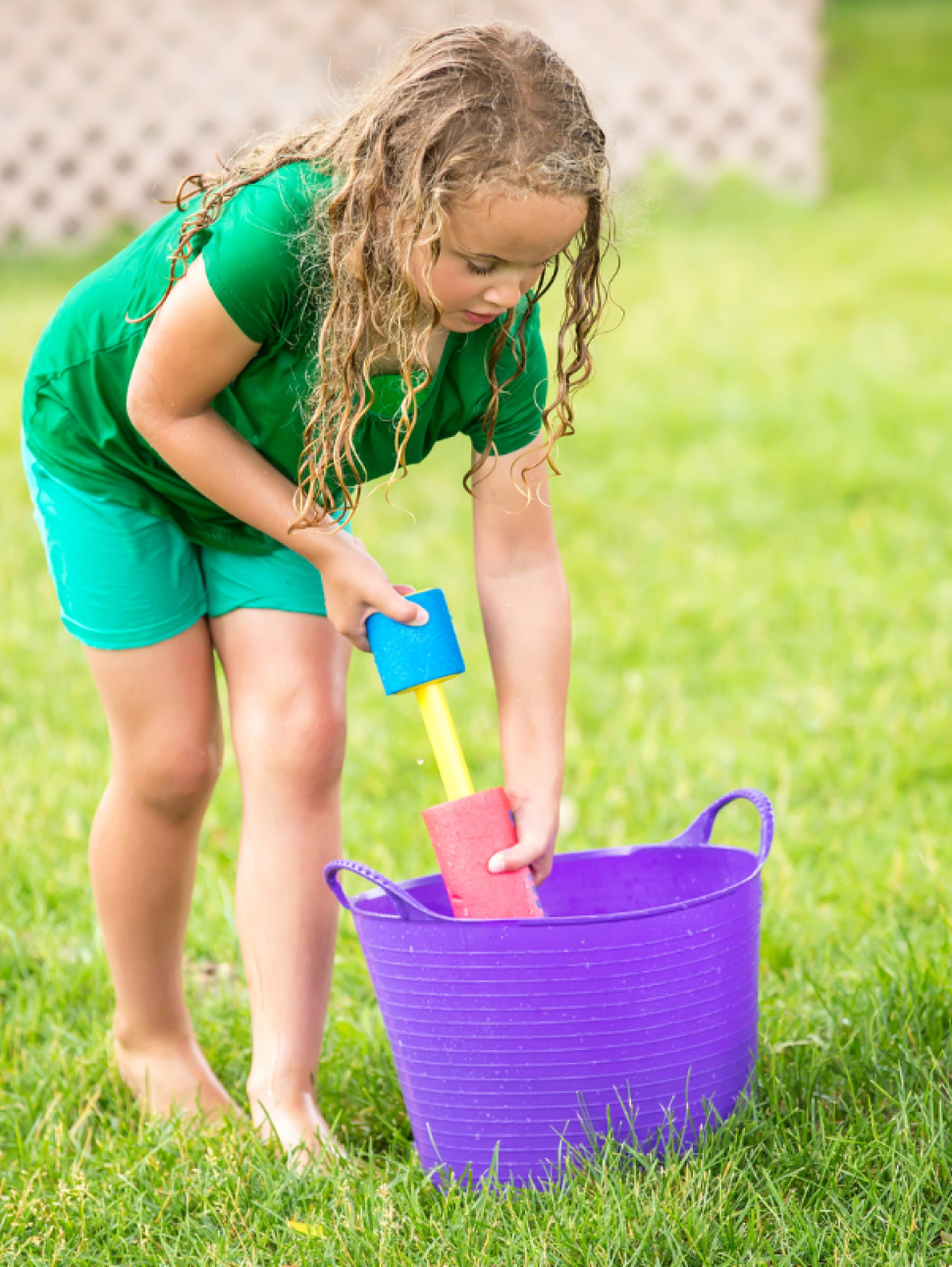 Child holding a water gun, drawing water from a bucket with green grass and a tree in the background