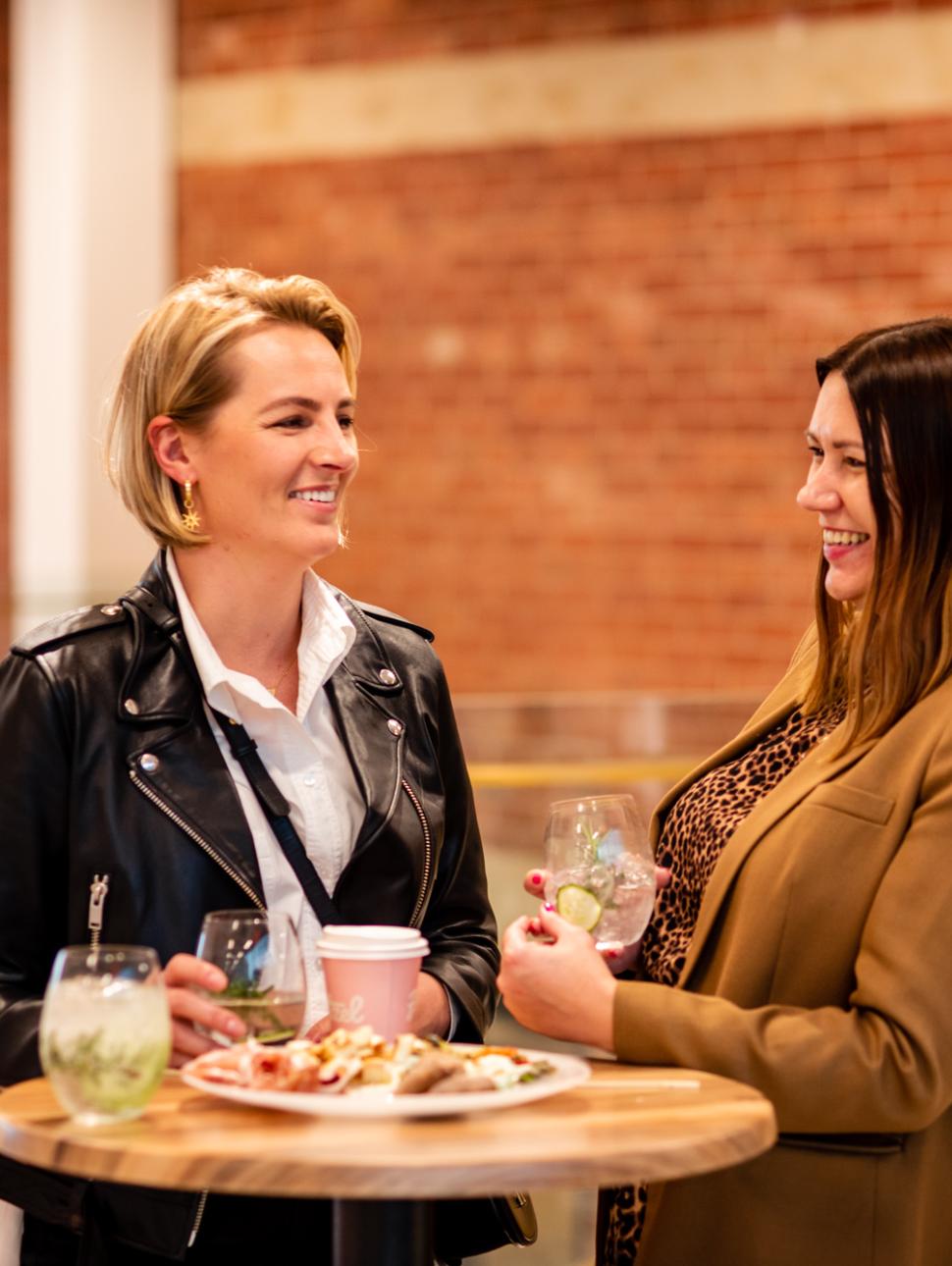 Two women stand over a high top table smiling, holding gin