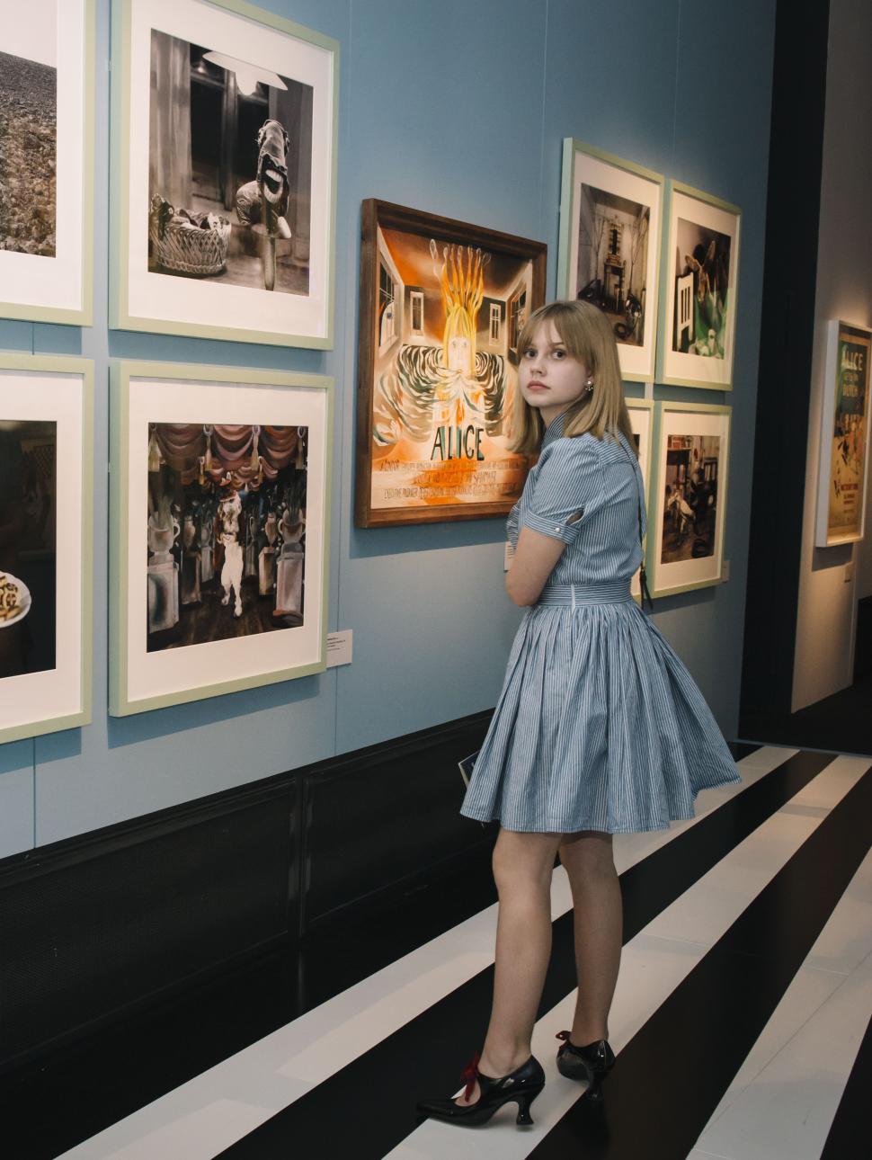 Girl dressed as Alice in Wonderland looks at a blue wall covered in Wonderland drawings and photos.