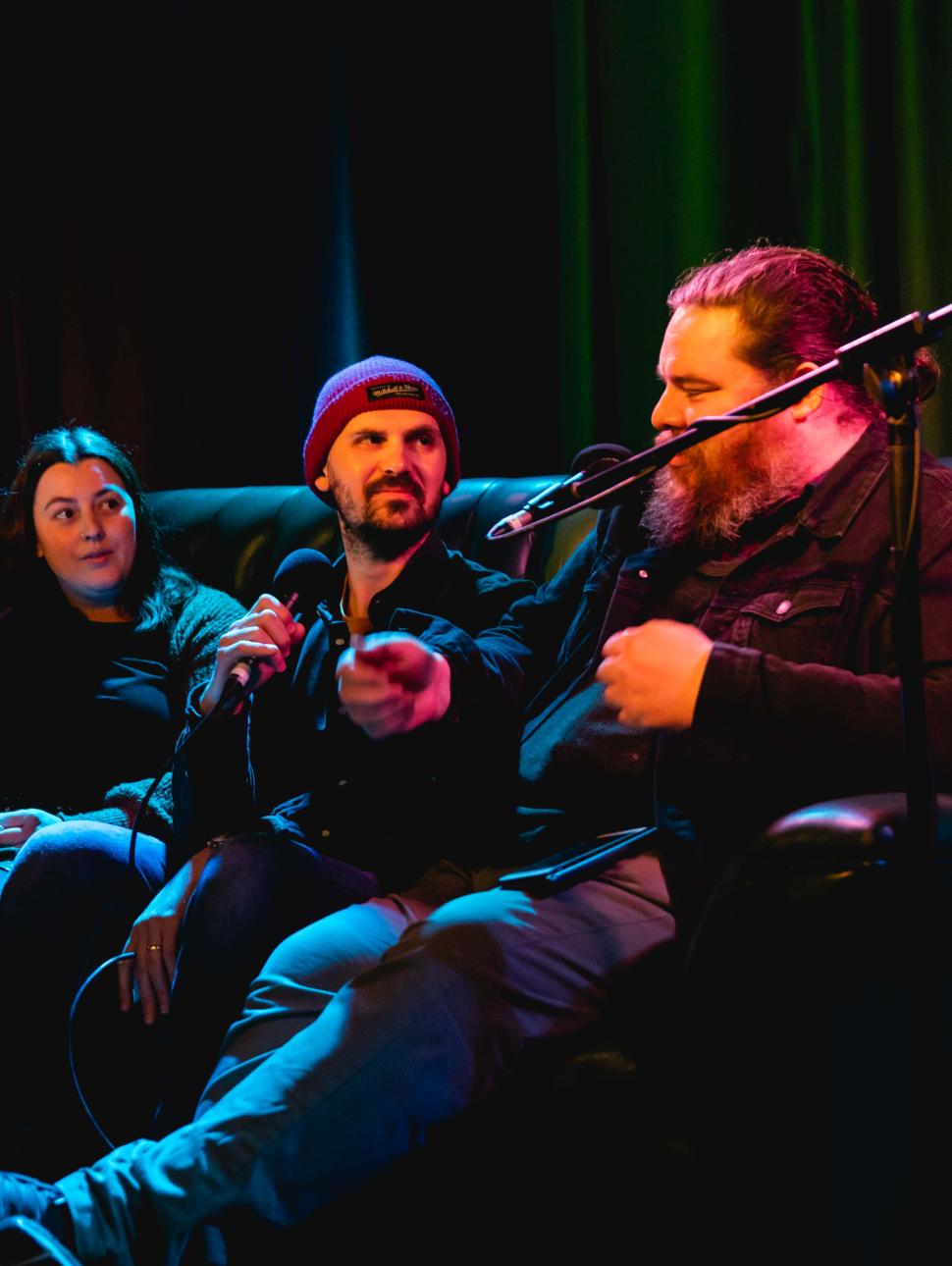 Three people sit in a dark room on a couch, speaking into microphones