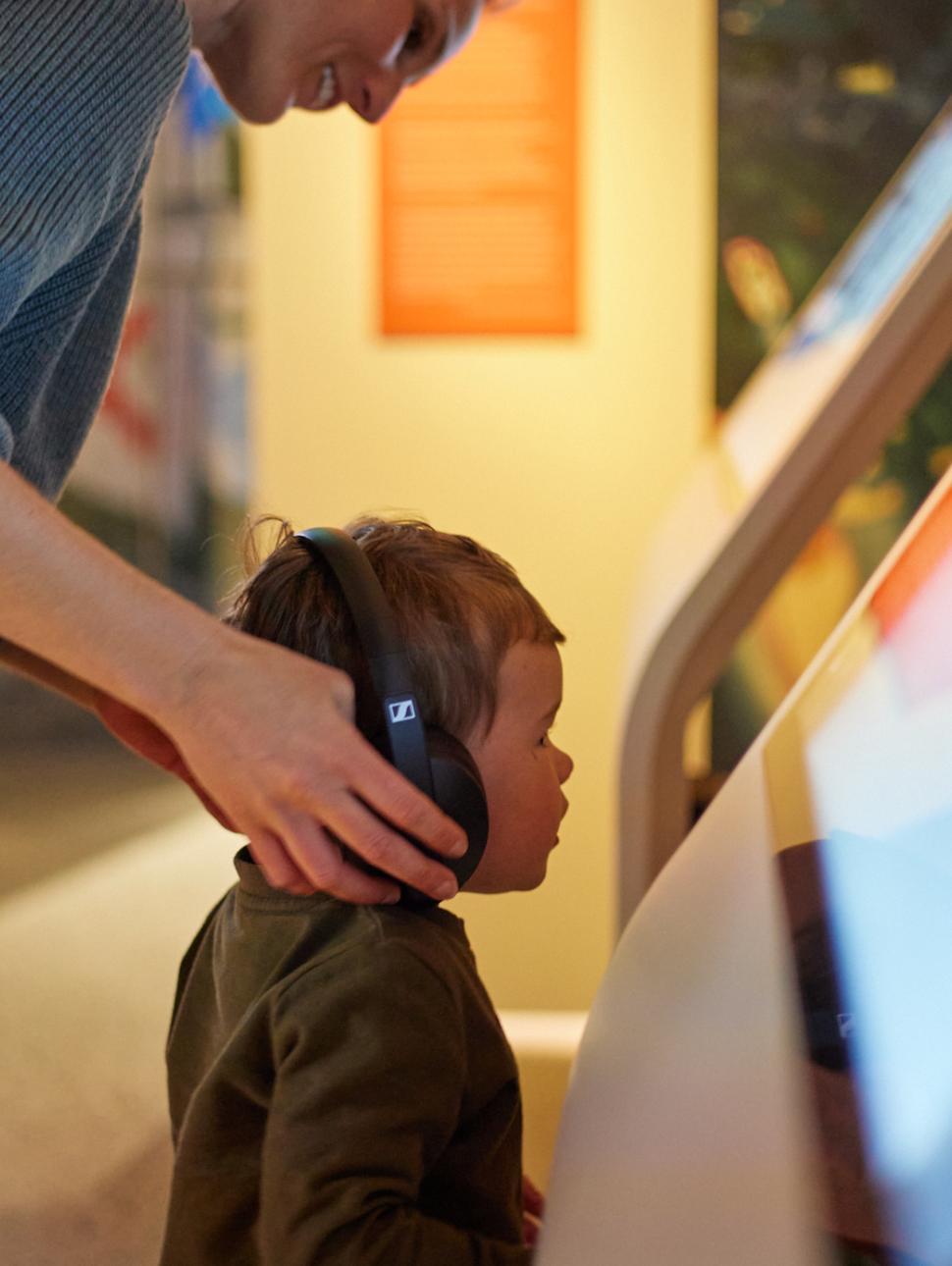 Image of a child wearing headphones looking at a digital screen with a parent behind 