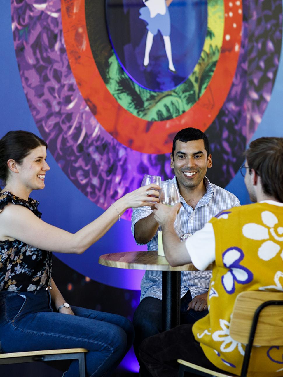 Three people are sitting at a high top table. One has their back to us. They have smiles on their faces and are in the middle of a cheers gesture with their drinks 