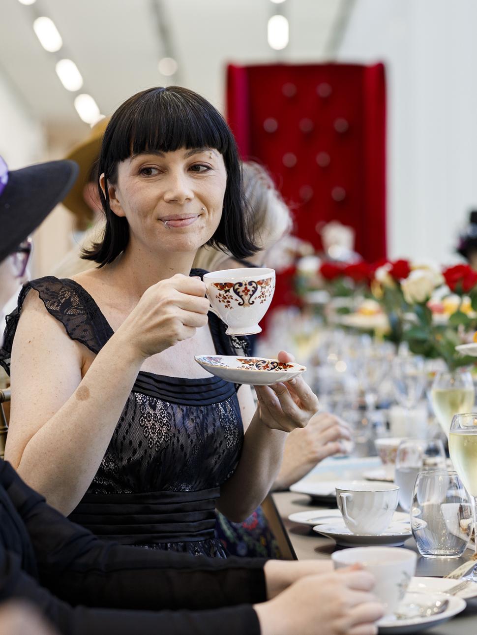People sitting at a long table holding teacups. The table is set with food and drink items