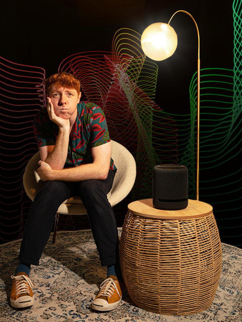 A person with a brown shirt, black jeans and orange sneakers sits on a sofa next to a light infront of LED lights