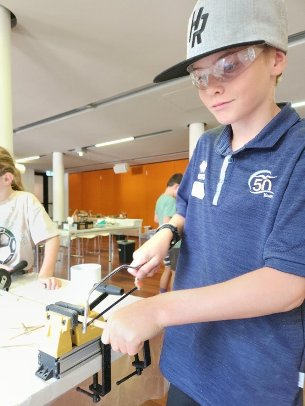 The image show a boy with using a hand saw and a vice to cut bamboo. He is waring afety glasses. 