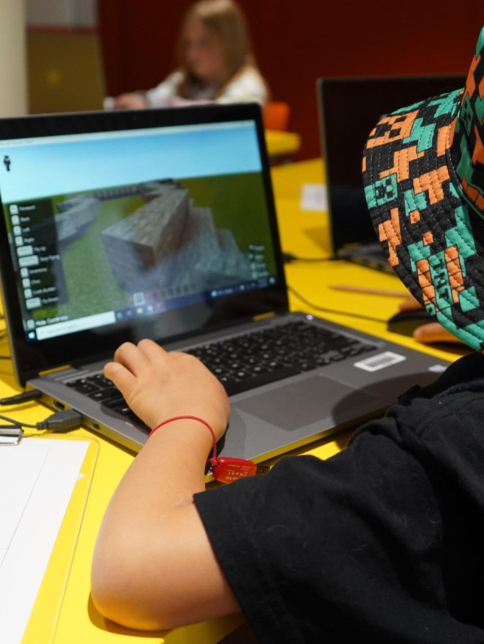 Image of a young person in a Minecraft hat plating Minecraft on a laptop.