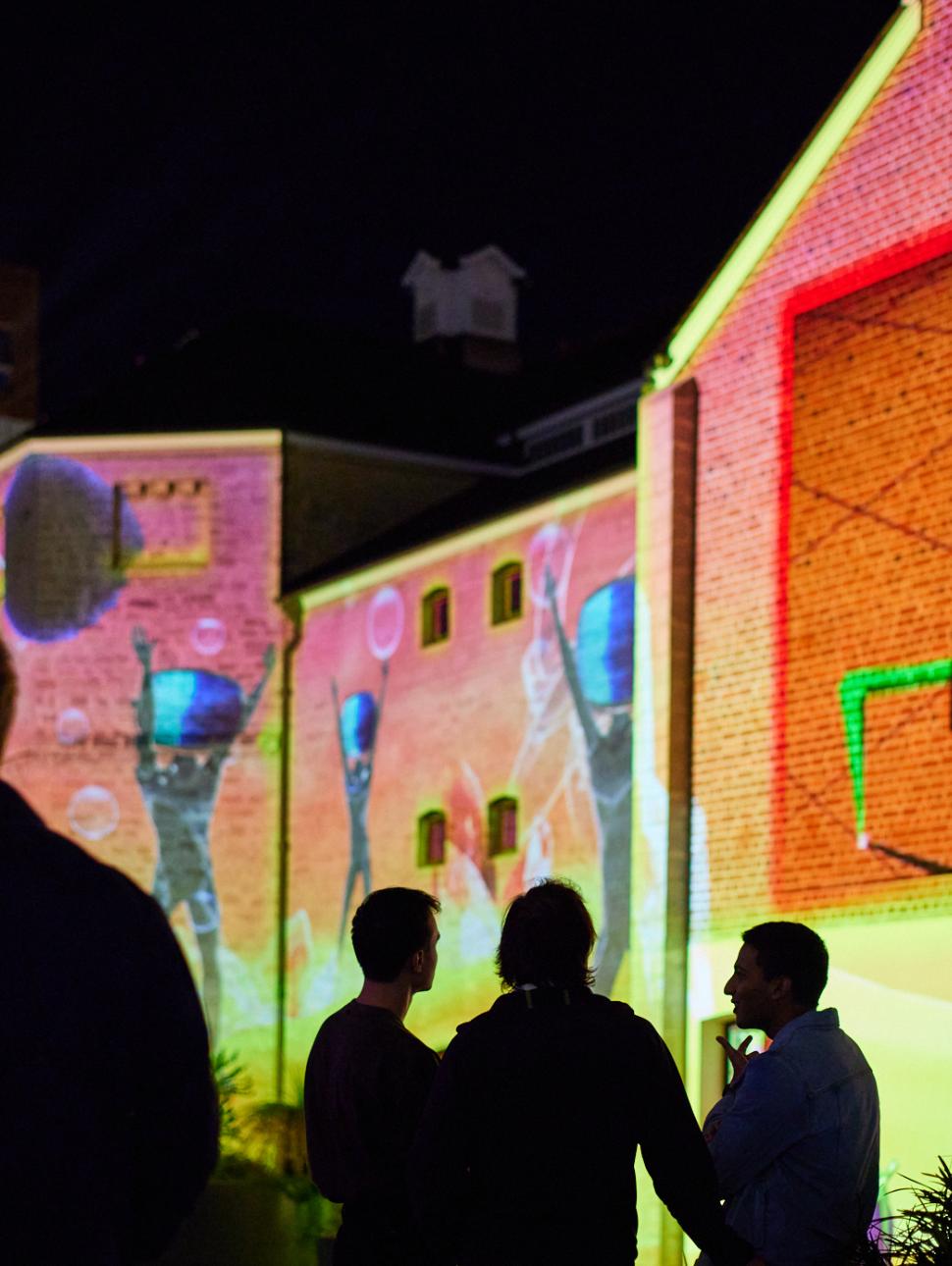 Colourful projections of Wonderland animations on the Old Gaol with people's shadows in the foreground. 