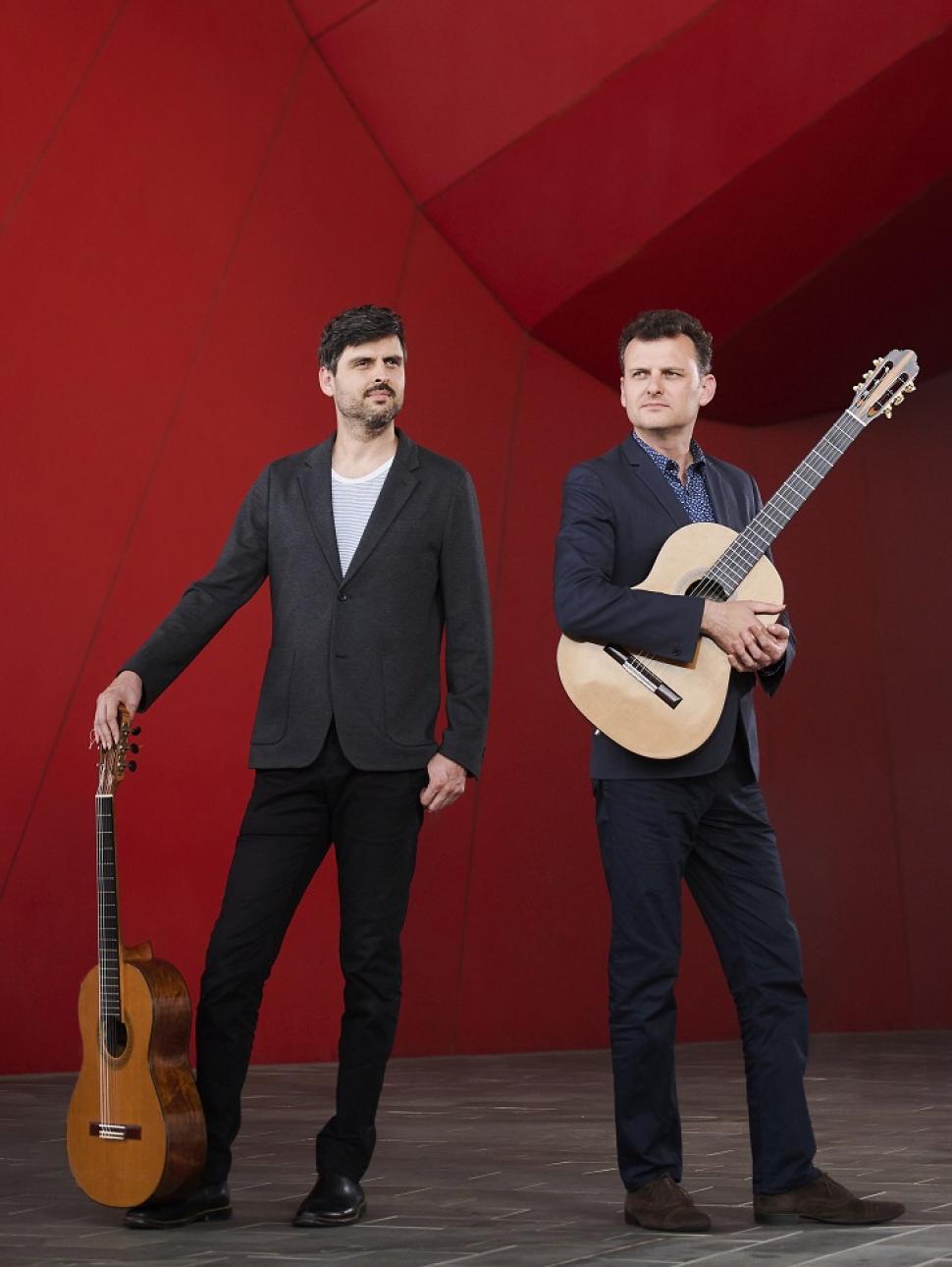 Two male presenting people stand with dark suits on and they hold guitars.