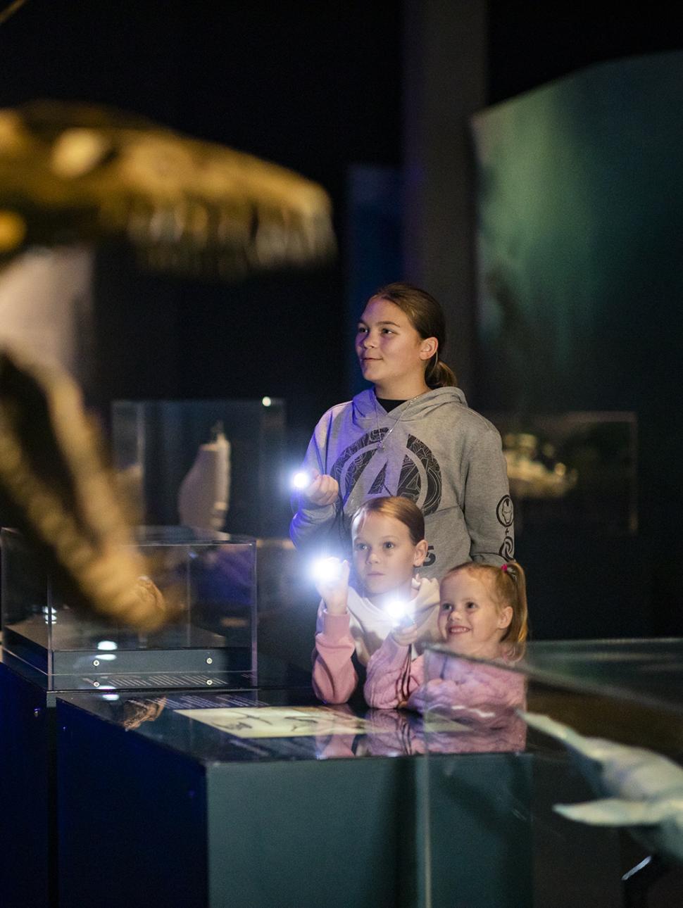 Three young people holding torches in a darkened exhibition with a skeleton in the foreground