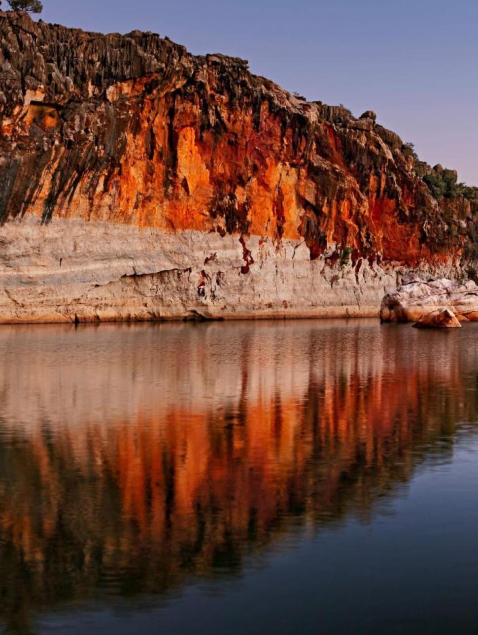 Orange, grey and sand coloured cliff reflected in the still water below with a clear sky above