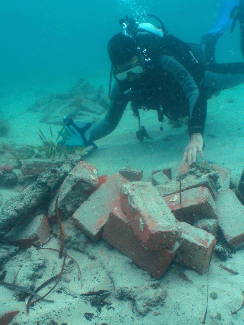 A scuba diver in full gear hovers over a pile of red bricks and pieces of wood at the bottom of the sea floor.
