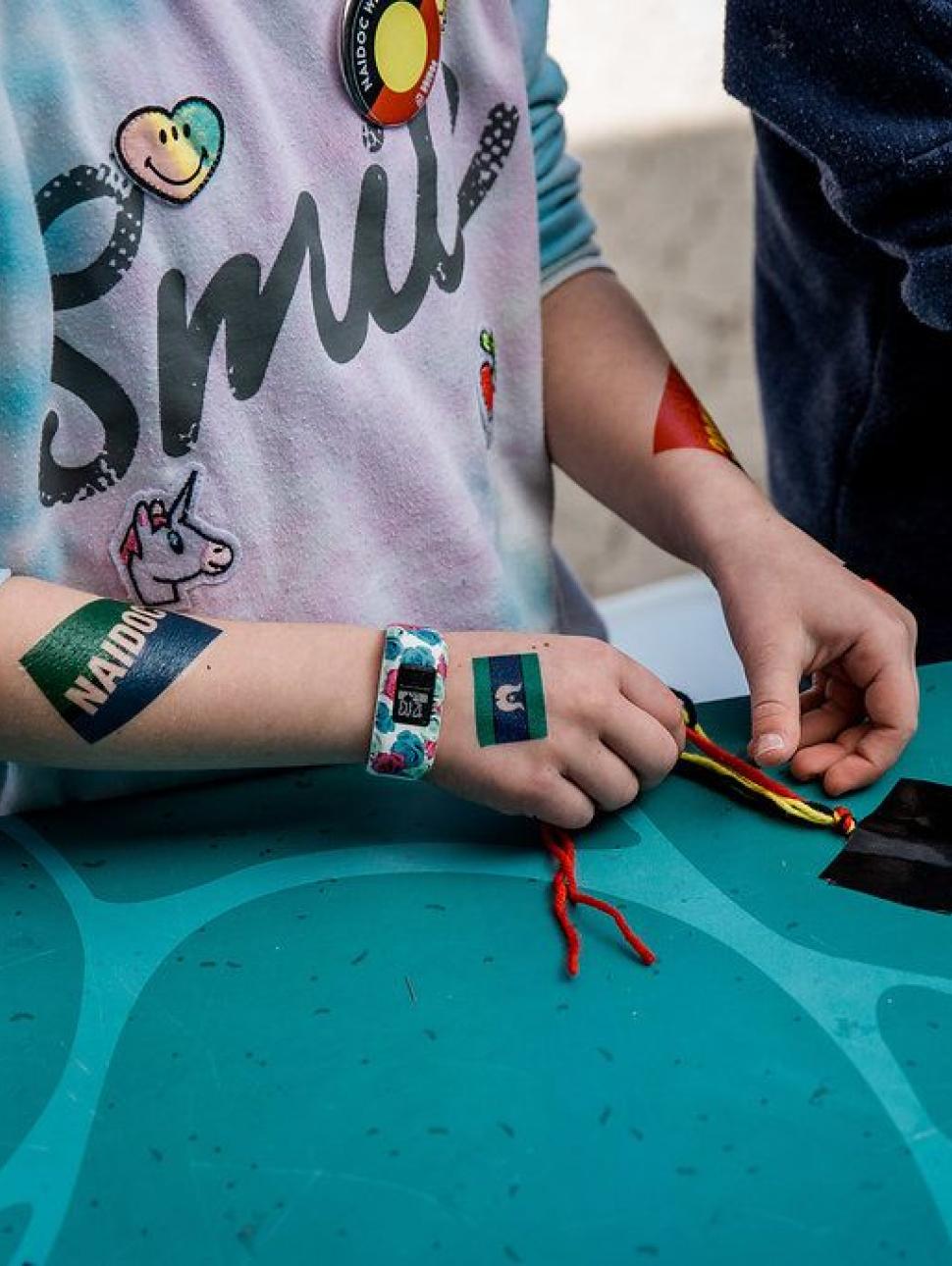 Image shows the hands of  child weaving a friendship bracelet from wall. They are working on a table top. 