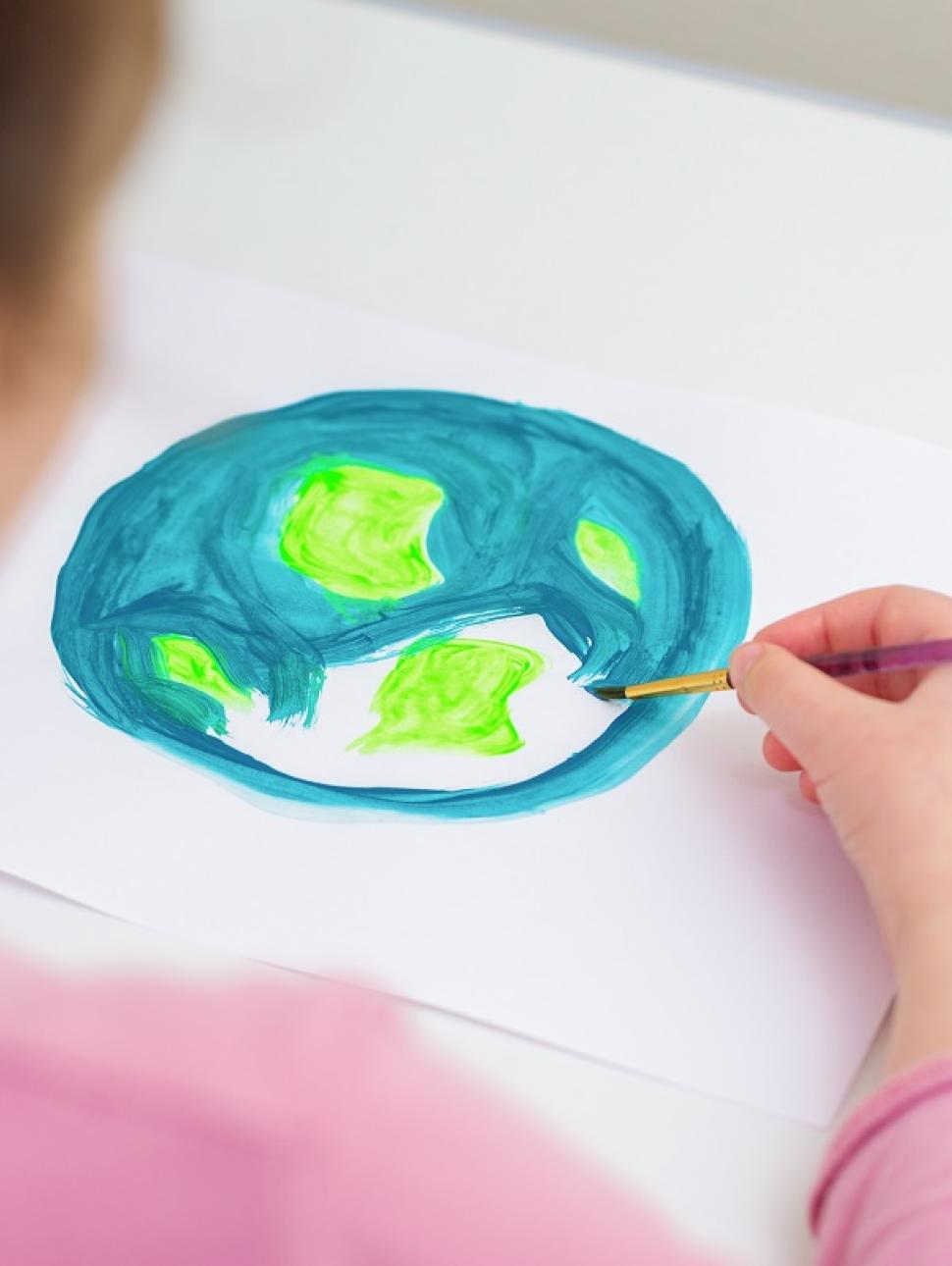 young child with back to the camera, focus of the image is of a painting on paper. They paint with blue and green, showing the earth. ocean and land.