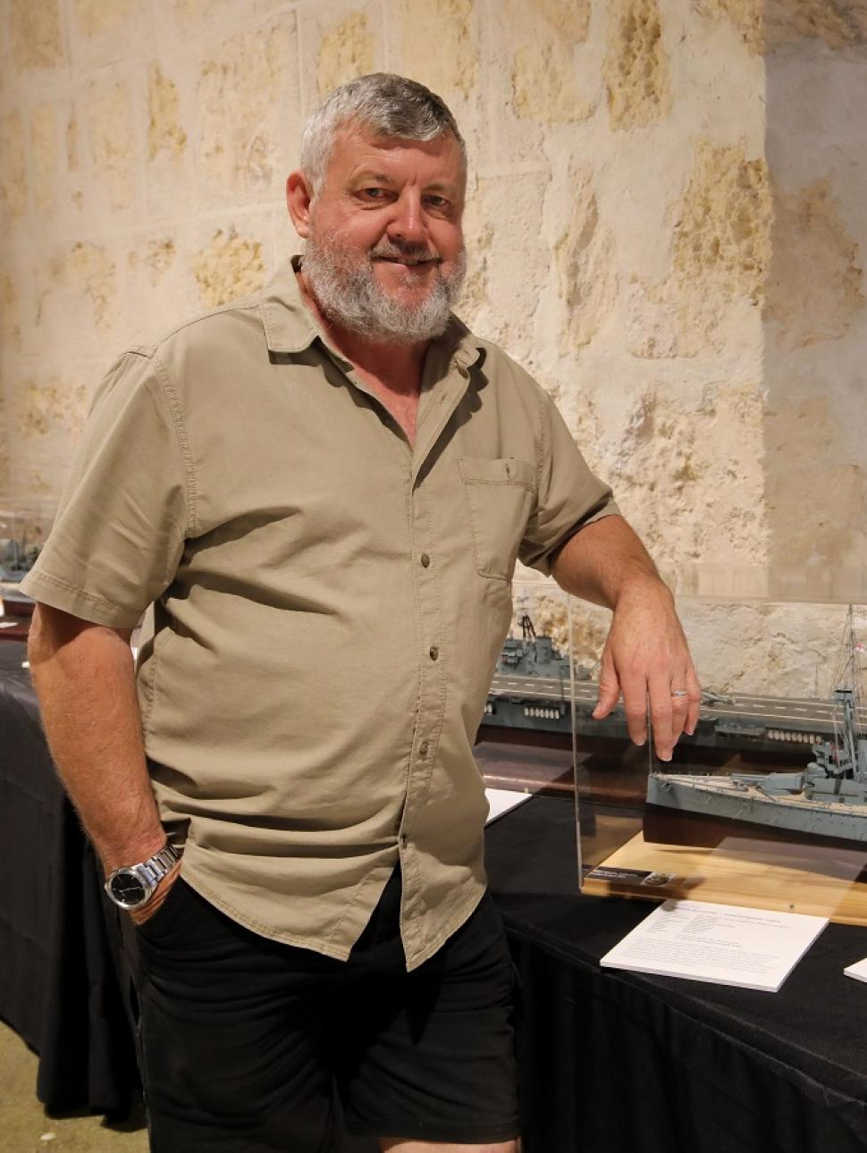 A person with a beard stands smiling in front of model warships display