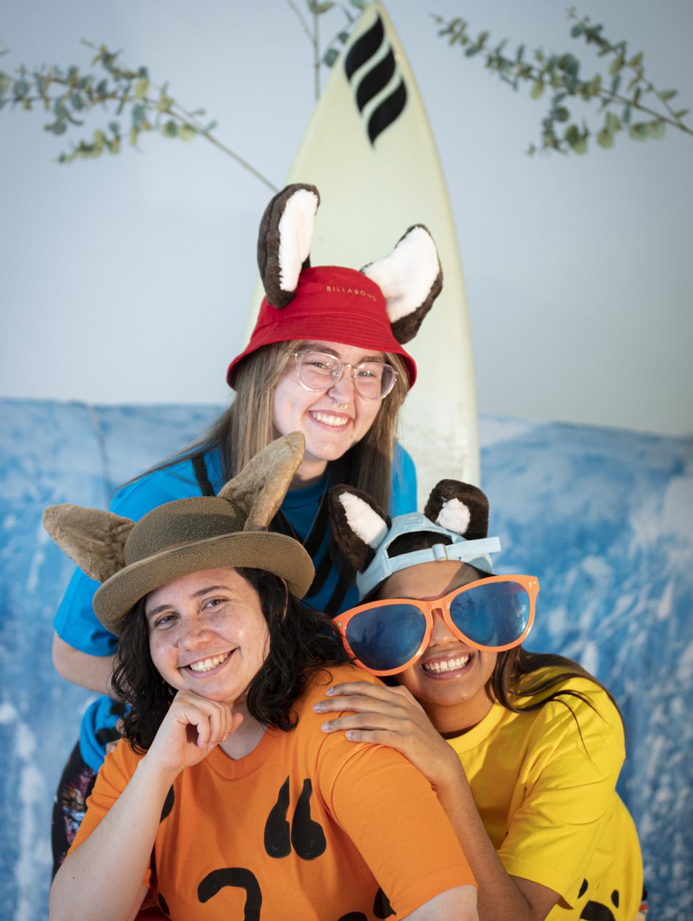 Three people are sitting smiling with bright coloured shirts on and headwear which mimics different animals.