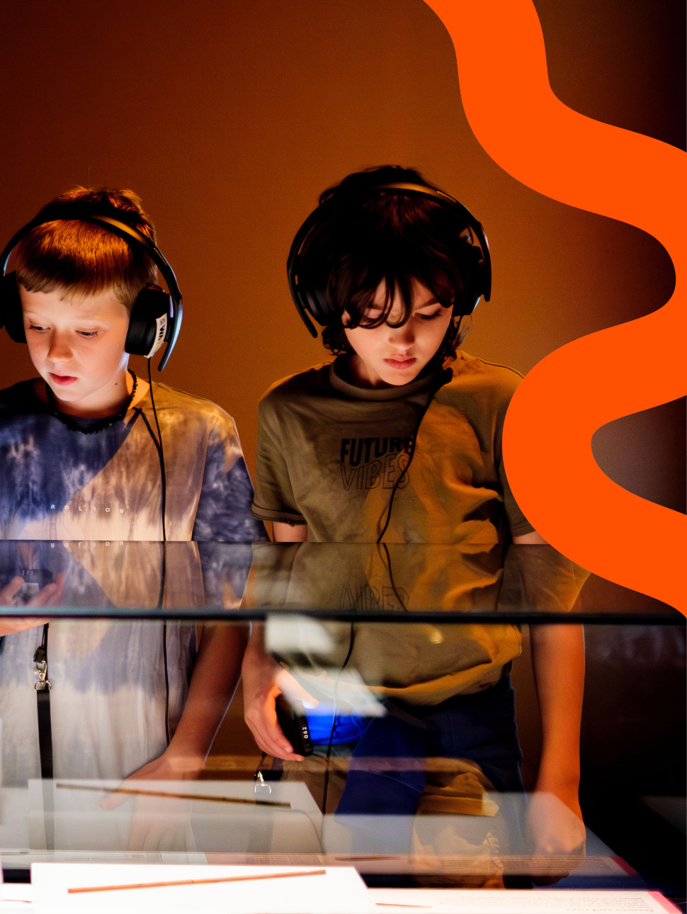 Two young kids wearing earphones, one with a brown top the other with a blue tie dye shirt. They are both looking down at a display cabinet.