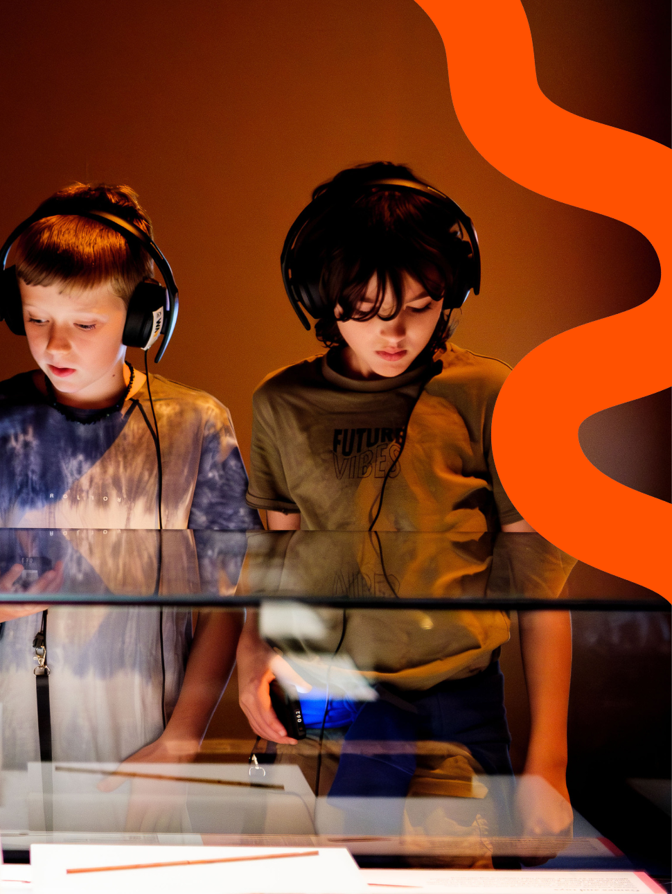 Two young kids wearing earphones, one with a brown top the other with a blue tie dye shirt. They are both looking down at a display cabinet. 