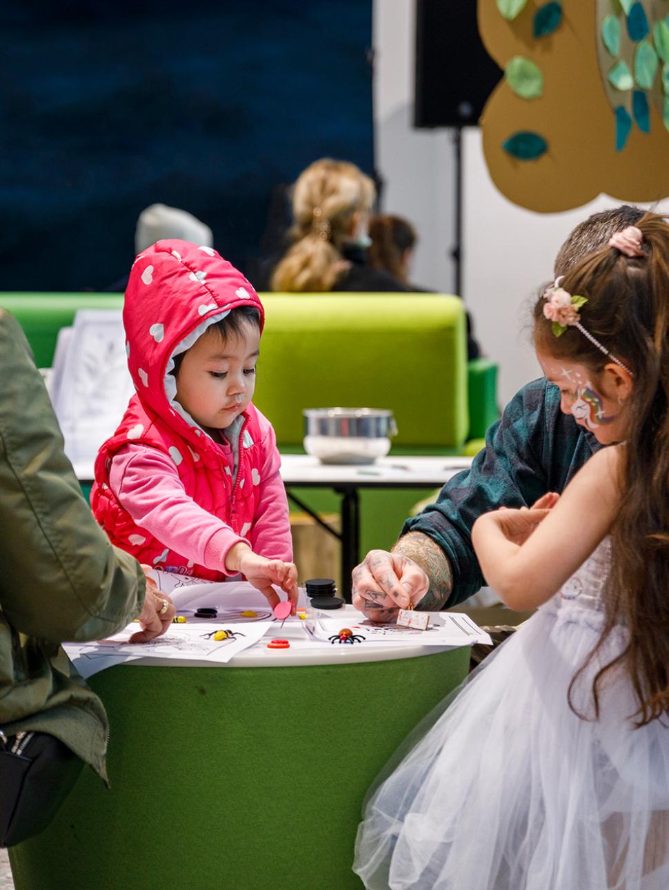 young children are crafting around a small round table with their adult carers. there are mini insects and magnets on the table